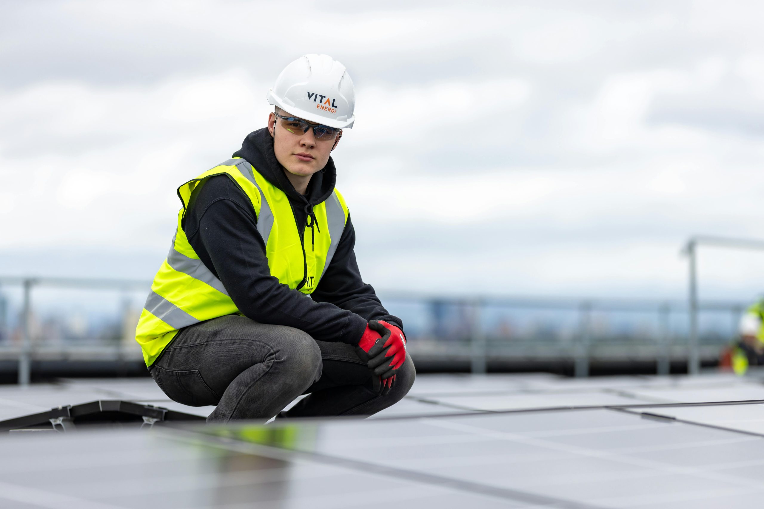 A construction worker inspects rooftop solar panels, wearing safety gear and high visibility vest.